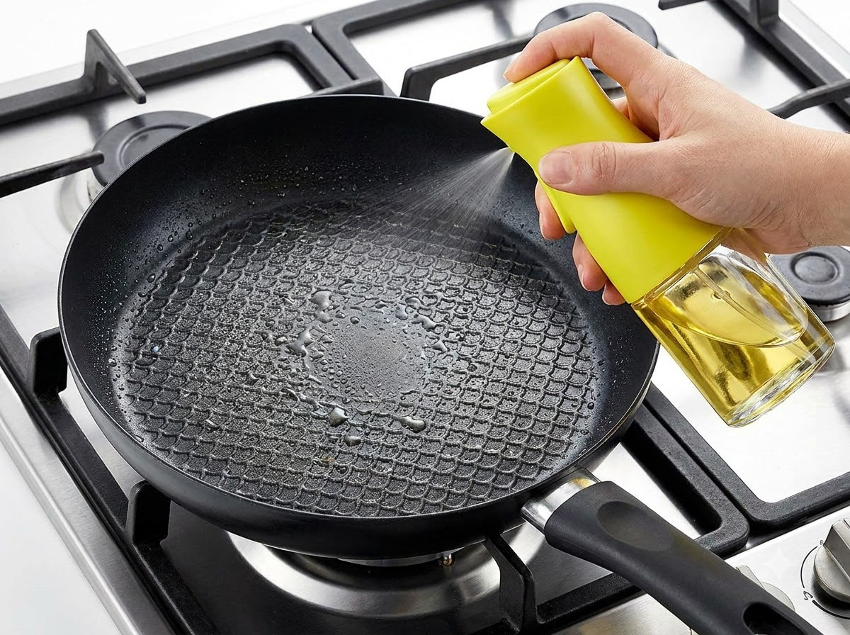 Une main vaporisant de lhuile de cuisson dans une poêle noire sur une gazinière.