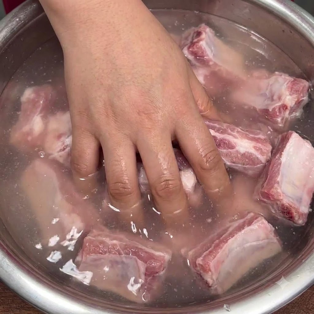 Une main mélangeant vigoureusement des côtes de porc crues dans un bol deau trouble pour répartir le bicarbonate.