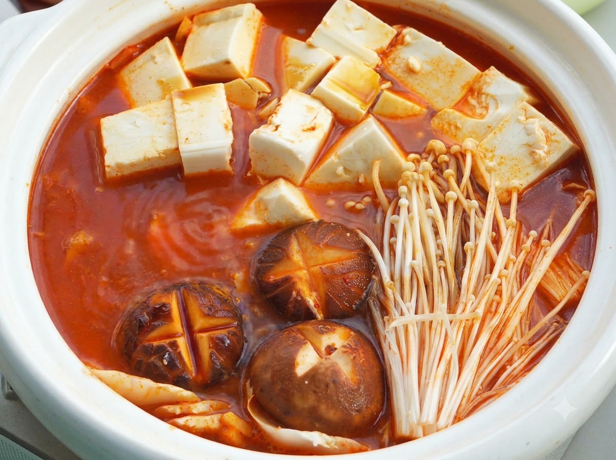 Une cocotte en céramique blanche remplie de base de soupe rouge, de cubes de tofu soyeux, de champignons shiitake incisés et de champignons enoki sous un couvercle.