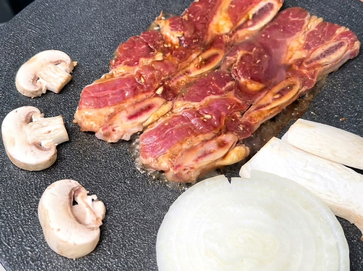 Travers de bœuf marinés grésillant sur une poêle à griller noire très chaude, à côté de tranches doignons blancs et de champignons.