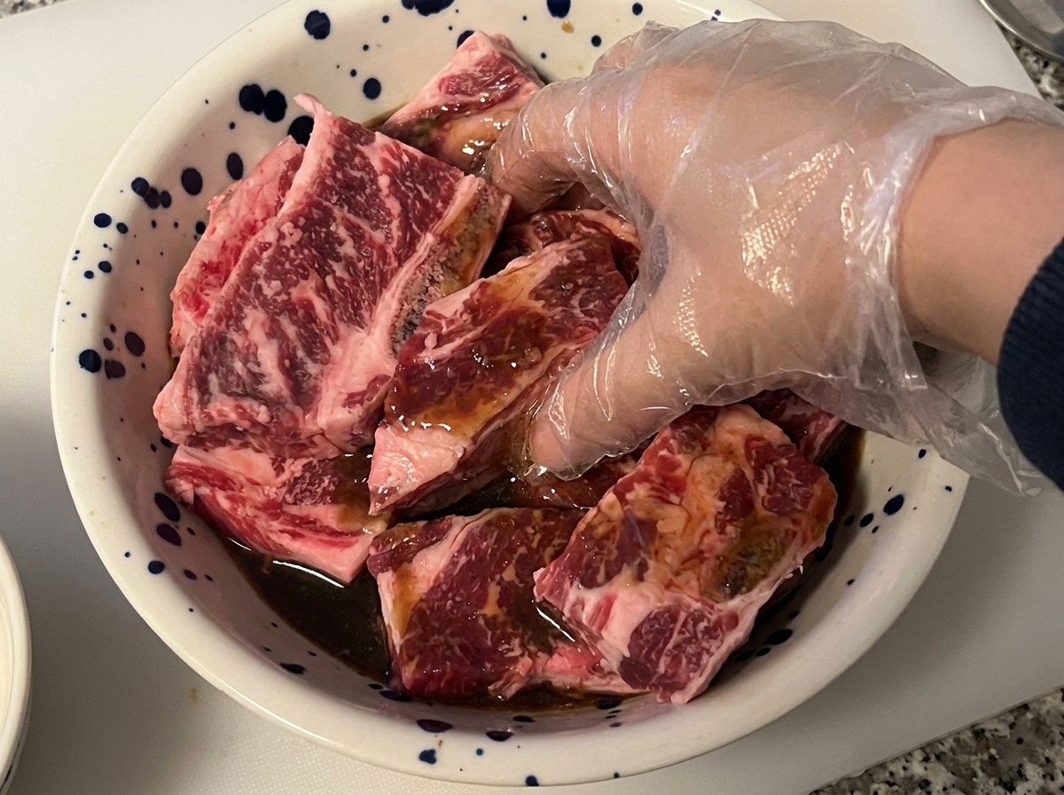 Une main gantée mélangeant et massant des côtes de bœuf crues avec une marinade foncée dans un bol.