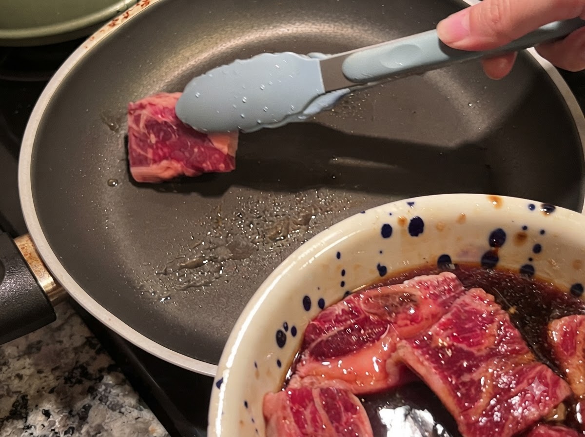 Des pinces plaçant un morceau de côte de bœuf marinée dans une poêle chaude légèrement huilée.