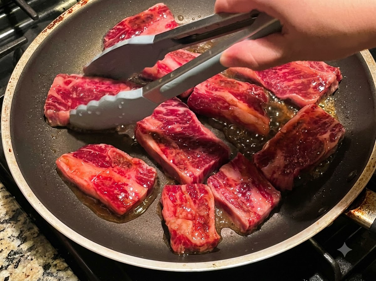 Des pinces retournant plusieurs morceaux de côtes de bœuf en train de frire dans une poêle.