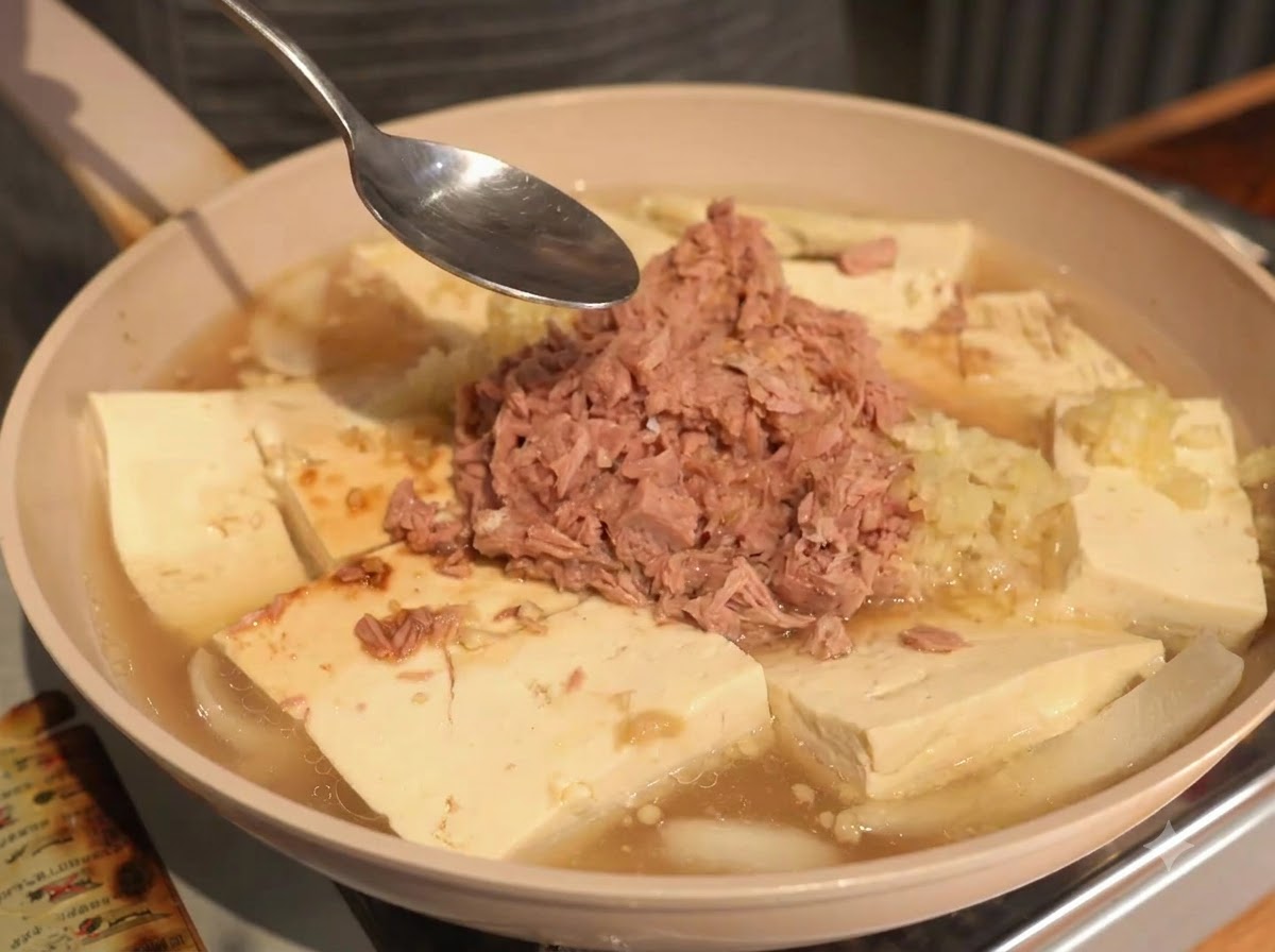Cuillère tenue au-dessus de la poêle contenant le thon, le tofu et le bouillon pendant lajout des poudres.