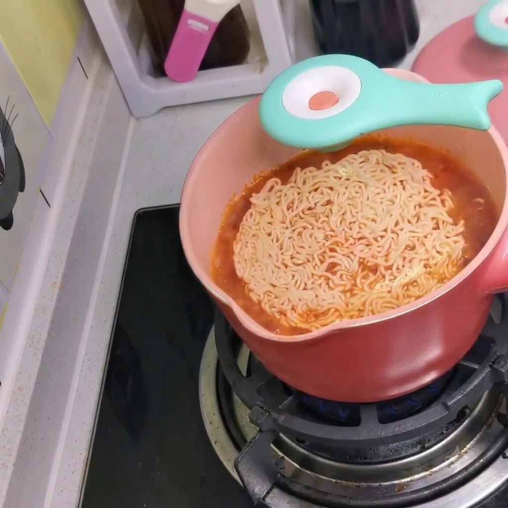 Un paquet de nouilles instantanées jeté dans une casserole rose de bouillon rouge picênt bouillant sur une cuisinière.