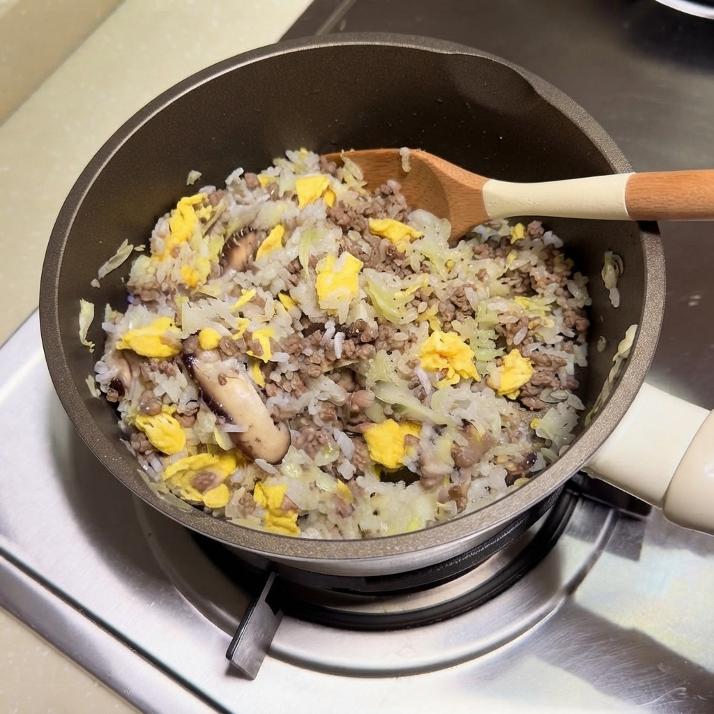 Un mélange de riz, de bœuf haché, de légumes et dœufs brouillés en train dêtre remué ensemble dans une cocotte avec une spatule en bois.