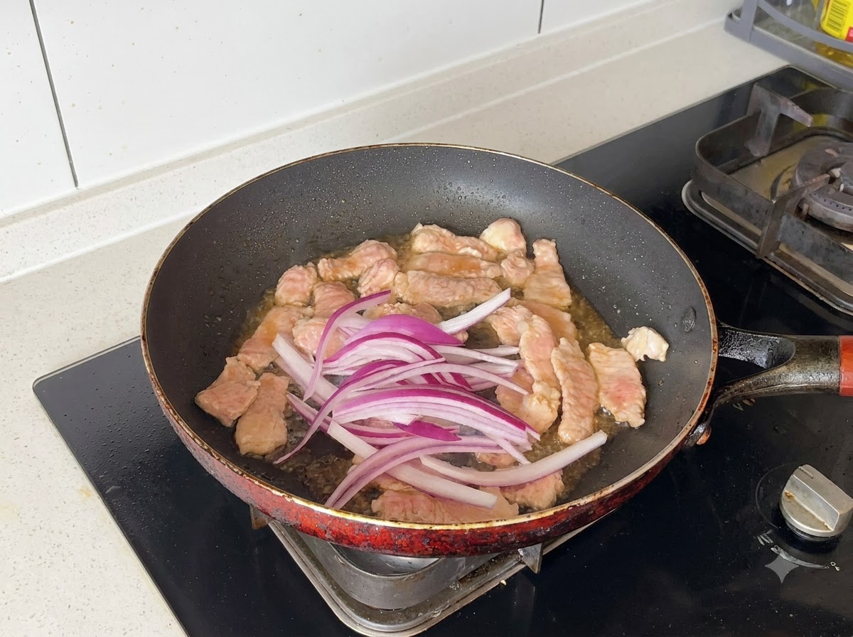 Des oignons rouges tranchés mélangés dans la poêle avec les morceaux de porc saisis.