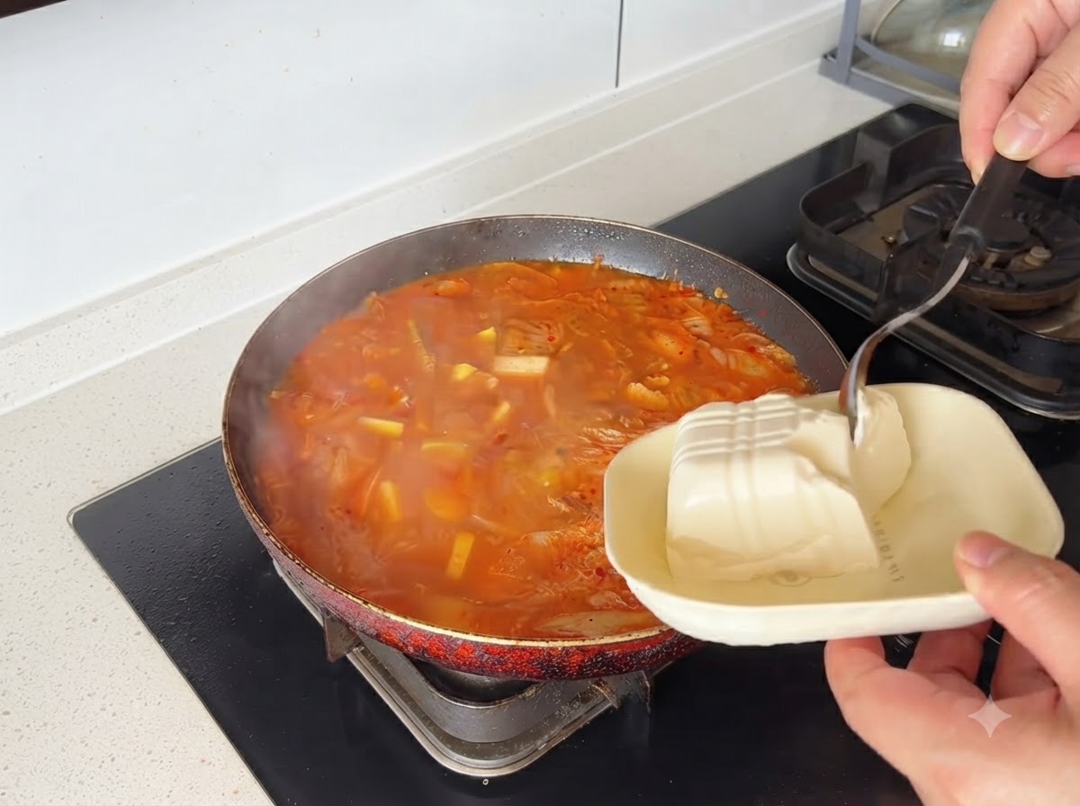 Des blocs de tofu blanc déposés dans une casserole de soupe au kimchi rouge.