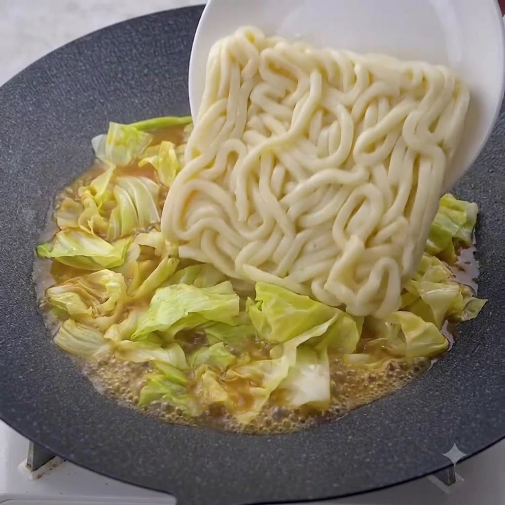 Un bloc de nouilles udon épaisses glissant dune assiette blanche vers une poêle contenant du chou et de la sauce frémissante.