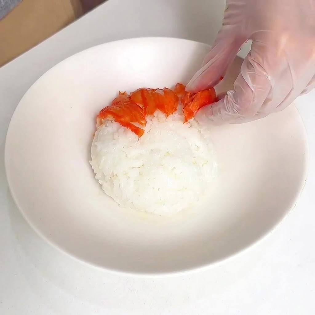 Une main gantée disposant des bâtonnets de surimi coupés en motif rayonnant sur un dôme de riz blanc dans un bol.